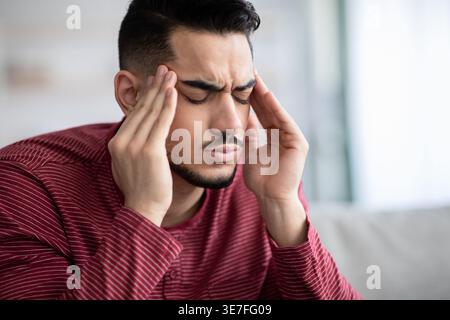 Middle Eastern man with closed eyes suffering from headache rubbing temples sitting on couch in living room at home Stock Photo