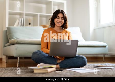 Smiling Hispanic woman studying using laptop sitting with crossed legs on floor with books and smartphone in front of bed in bedroom at home Stock Photo