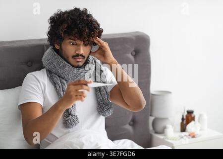 Sick Asian man with curly hair wearing scarf around his neck touching head and holding thermometer checking body temperature while sitting on bed i... Stock Photo