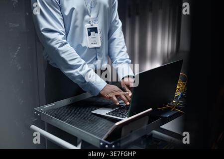 Black male network engineer using laptop and setting up servers while standing in data center Stock Photo