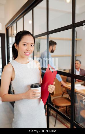 Smiling Asian business woman holding cup of coffee and documents folders standing in office Stock Photo