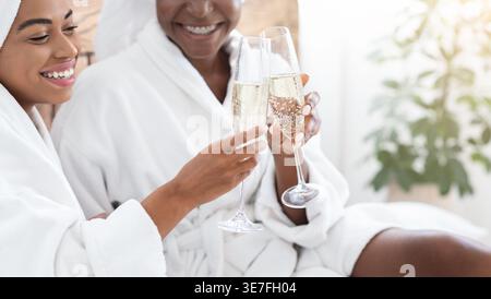 Smiling Black women wearing bathrobes clinking glasses of champagne while sitting on bed in bedroom at home Stock Photo