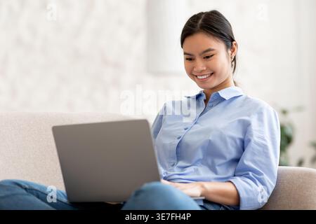 Smiling Asian woman using laptop sitting on couch with crossed legs in living room at home Stock Photo