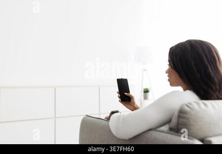 Black woman using smartphone sitting on sofa in living room at home Stock Photo