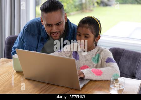 Father and child sitting at wooden dining table using silver laptop with yellow headband and mug Stock Photo