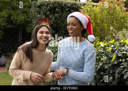 Diverse female friends standing close holding hands in garden wearing Santa hat and headband Stock Photo