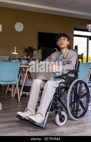 Asian man sitting in wheelchair, wearing denim jacket, using silver laptop in coworking lounge Stock Photo