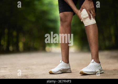 Black man wearing sportswear wrapping injured knee while standing outdoors during daytime Stock Photo