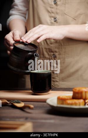 Asian woman pouring tea in ceramic cup while standing at table with mooncakes indoors Stock Photo