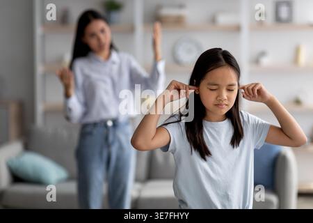 Asian girl with closed eyes covering ears with fingers while standing near mother scolding her in living room at home Stock Photo