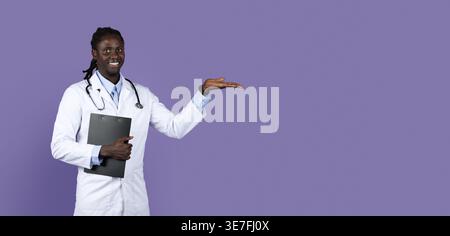 Smiling Black male doctor with dreads wearing white suit with stethoscope holding clipboard showing aside while standing against purple background ... Stock Photo