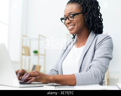 Smiling Black business woman with twists wearing gray coat and eyeglasses using laptop while sitting at table in office Stock Photo