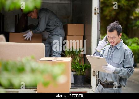 Asian man having phone call and checking document standing near male coworker loading boxes into truck outdoors during the daytime Stock Photo