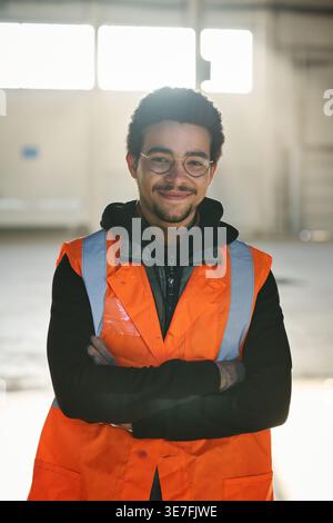 Mixed-race male worker wearing eyeglasses and safety vest standing with arms crossed in warehouse Stock Photo