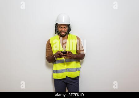 Black male engineer with twists wearing hard hat and safety vest using smLifestylephone while standing against white wall Stock Photo