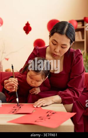 Asian mother and daughter writing with ink and brush doing traditional calligraphy while sitting in living room at home Stock Photo
