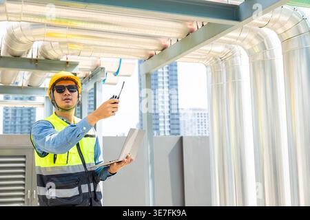 Asian male engineer holding laptop and walkie talkie checking pipelines standing on rooftop during daytime Stock Photo
