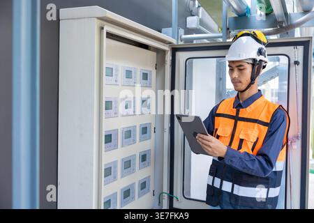 Asian male worker wearing safety helmet using tablet checking main circuit cabinet switchboard control standing on rooftop during daytime Stock Photo