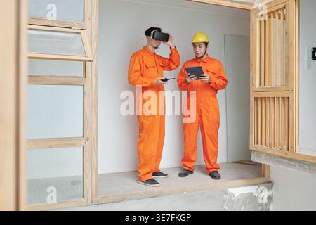 Mixed race male construction worker using virtual reality headset talking to Asian male construction worker holding tablet while standing together ... Stock Photo