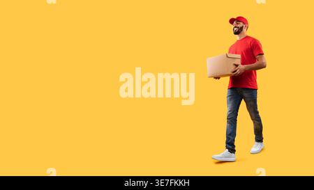 Arab delivery man wearing cap holding cardboard box walking against yellow background in studio Stock Photo