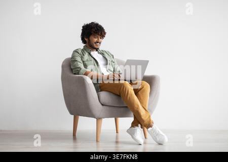 Asian man with curly hair using laptop while sitting on chair against white wall Stock Photo