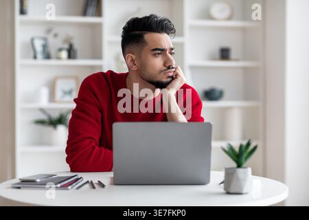 Bored Arab man looking away leaning chin on hand sitting at desk with laptop at home Stock Photo
