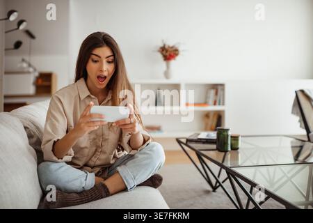 Shocked Hispanic woman playing games on her smartphone while sitting with crossed legs on sofa in living room at home Stock Photo