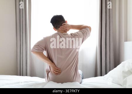 Asian man suffering from neck and lower back pain sitting on bed near window in bedroom at home Stock Photo