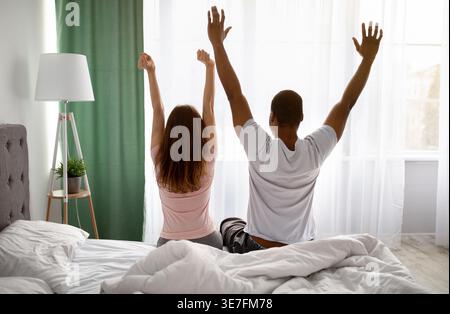 Black and caucasian couple waking up stretching their arms while sitting on bed at home Stock Photo