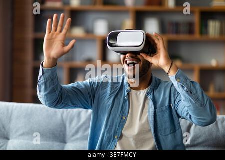 Black man reaching hand forward wearing VR Glasses enjoying virtual reality experience sitting on sofa in living room at home                      ... Stock Photo