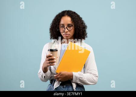 Tired Black female teenager wearing eyeglasses holding books and cup of coffee while standing against blue background in studio Stock Photo