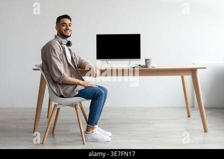 Smiling Middle Eastern man with headphones on neck sitting at table with computer and coffee cup in office Stock Photo