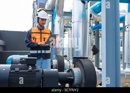 Asian male worker wearing safety helmet using tablet checking pipeline system standing on rooftop during daytime Stock Photo