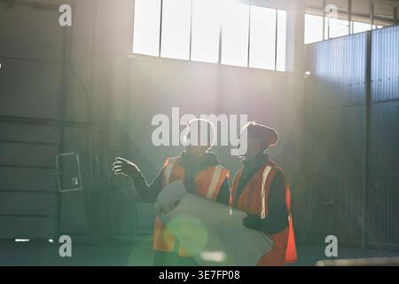 Mixed-race male engineers wearing safety vests talking with each other holding blueprints standing together in warehouse with sunlight Stock Photo