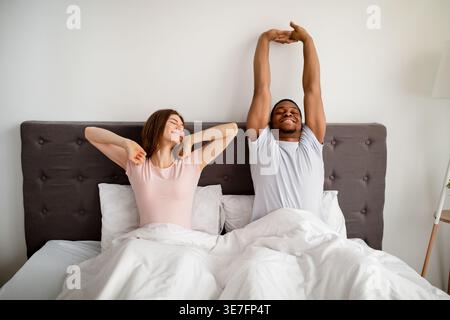 Black and caucasian couple waking up stretching their arms while sitting on bed at home Stock Photo