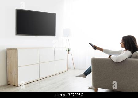 Black woman holding remote control and switching channels on TV with blank black screen, sitting on sofa in living room at home Stock Photo