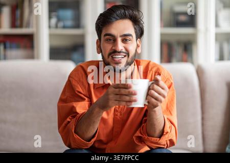 Smiling Middle Eastern male holding coffee cup sitting on sofa at home Stock Photo
