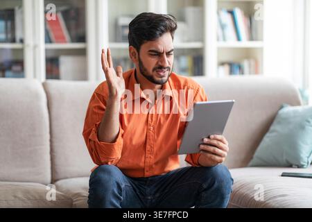 Upset Middle Eastern man raising hand having video call using tablet sitting on couch at home Stock Photo