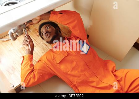 Black male plumber fixing pipe while lying under sink in kitchen of customer Stock Photo