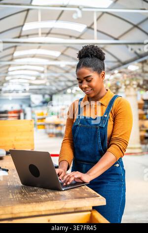 Hispanic female employee managing orders using laptop while standing at wooden table in garden center Stock Photo