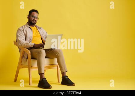 Serious Asian man with eyeglasses using laptop sitting on chair against yellow background in studio Stock Photo