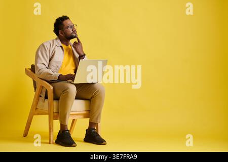 Confused Asian man with eyeglasses pointing finger on chin looking away sitting on chair with laptop against yellow background in studio Stock Photo