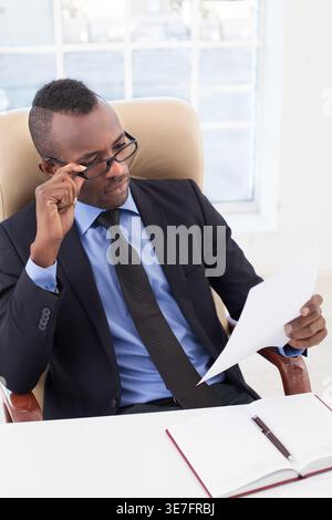Black businessman adjusting eyeglasses reading document sitting at table with notebook in office Stock Photo