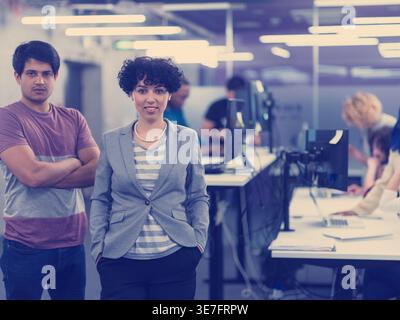 Smiling Mixed race woman with hands in pockets standing near Mixed race man with arms crossed in office Stock Photo