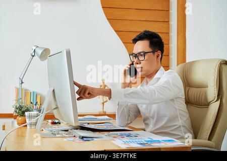 Asian male graphic designer talking on smartphone and looking at monitor screen while sitting on chair in office Stock Photo