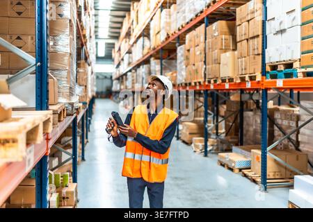 Black male worker with dreads wearing safety vest and hard hat scanning package codes using wireless scanner while standing in logistic warehouse Stock Photo
