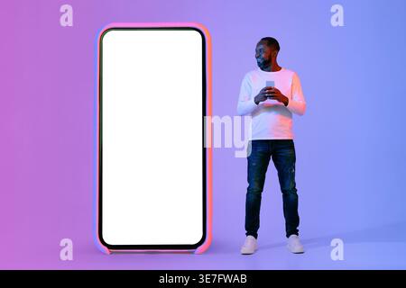 Smiling Black man using smartphone looking at huge phone while standing against purple background in studio with neon lights Stock Photo