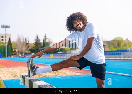 Black male runner with curly afro stretching leg on barrier standing on running track on sunny day Stock Photo