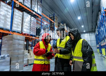 Diverse group of warehouse workers wearing safety vests and warm clothing using barcode scanner and clipboard while standing and inspecting product... Stock Photo