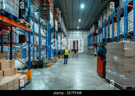 Hispanic male worker wearing black winter jacket and safety vest inspecting goods on high racks with male colleague driving forklift during work in... Stock Photo